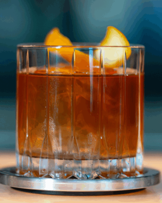 Close up photo of an old fashioned cocktail in a ridged crystal rocks glass that is sitting on a stainless steel coaster. The whiskey is over a large block cube of ice with an orange twist garnish on top.