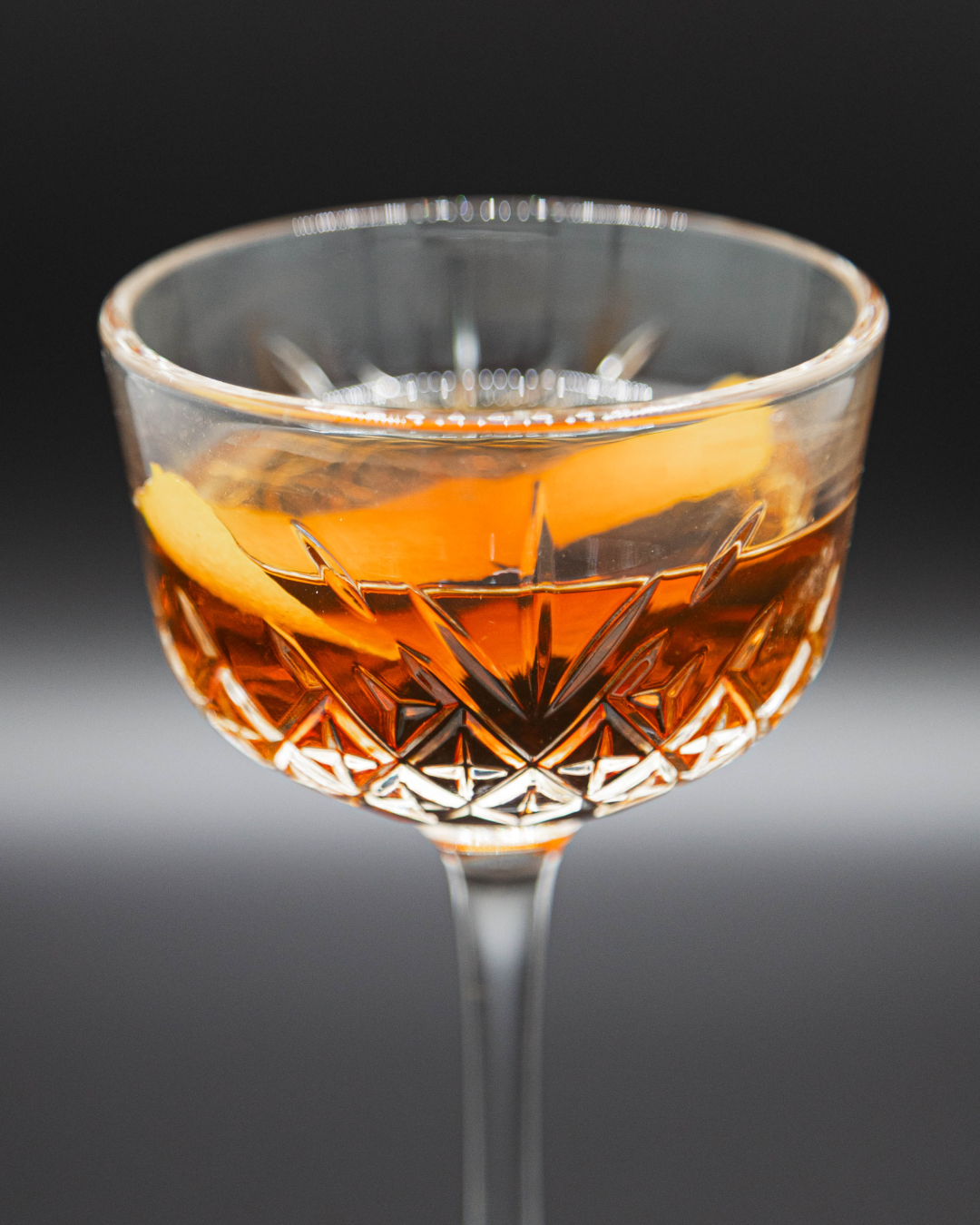 A tiny tini version of the Manhattan cocktail in a smaller crystal coupe glass with an orange peel garnish in the drink