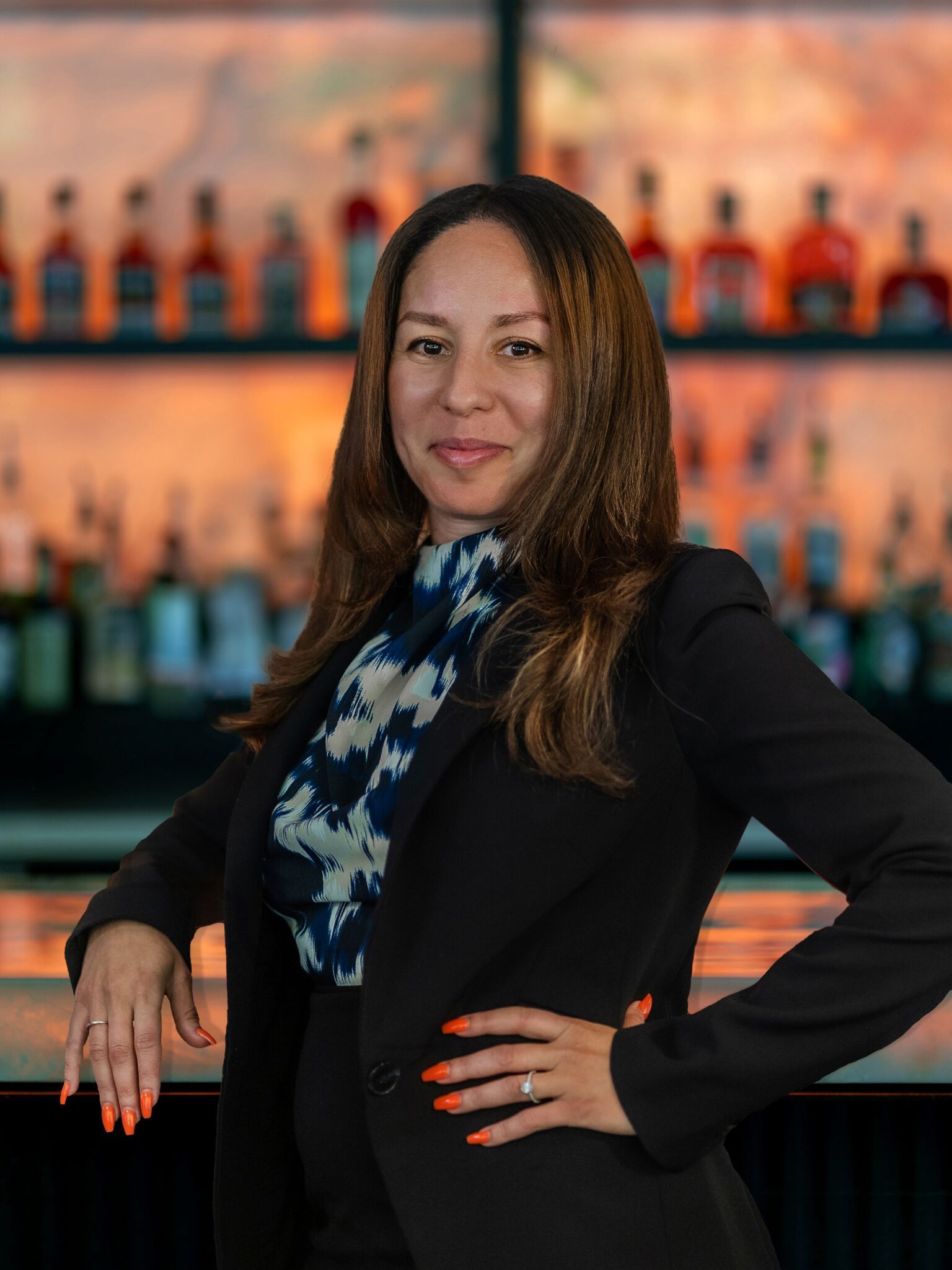 headshot of Austin Arboretum GM, Myriam Chiny. She is standing in front of the bar top with one hand on her hit and resting her other arm on the bartop. She has a dark suit on with a navy and tan print blouse underneath.
