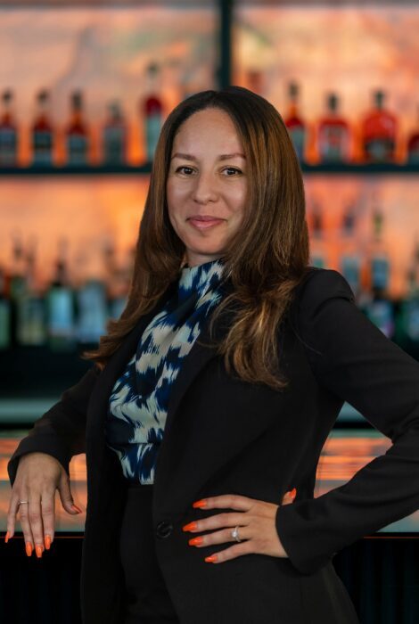 headshot of Austin Arboretum GM, Myriam Chiny. She is standing in front of the bar top with one hand on her hit and resting her other arm on the bartop. She has a dark suit on with a navy and tan print blouse underneath.