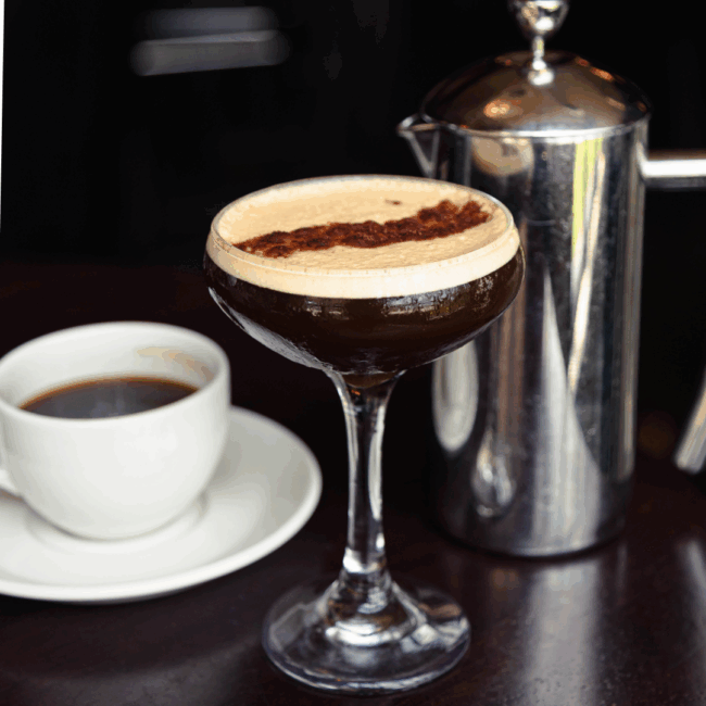 an image of an espresso martini in a coupe glass on the bar top with a cup of coffee on a white saucer and a stainless steel french press in the background