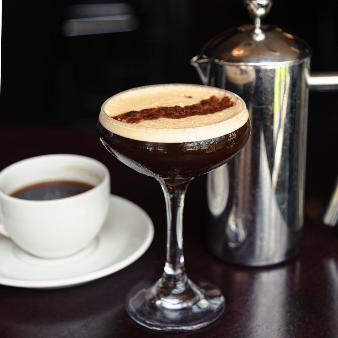 an image of an espresso martini in a coupe glass on the bar top with a cup of coffee on a white saucer and a stainless steel french press in the background