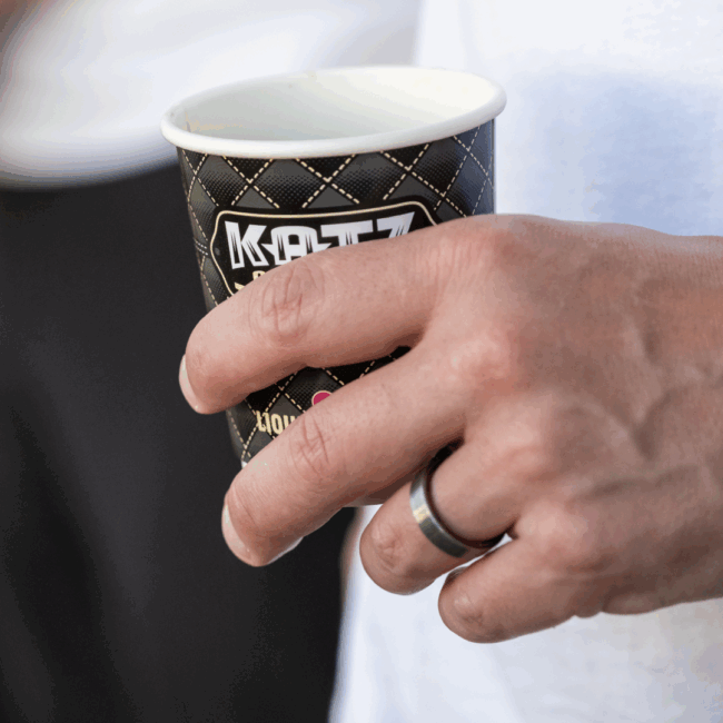 image of a male hand with a silver ring on the ring finger holding a paper coffee cup that says Katz on it
