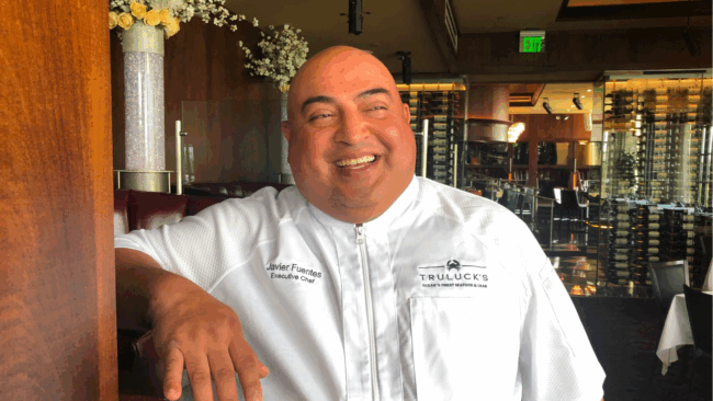 Chef Javier Feuntes smiling and laughing in a white chef coat with his arm propped on a booth in the dining room