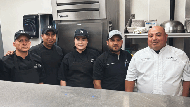 Chef Javier fuentes in a white chef coat standing next to his culinary team in the kitchen all dressed in black chef coats and Truluck's crab logo hats