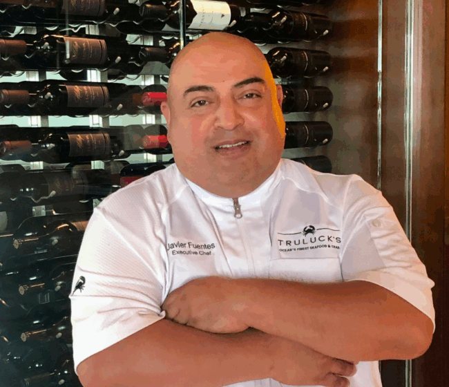 chef Javier Fuentes of Truluck's Rosemont in a white logoed chef coat standing in front of the wine wall with his arms folded across his chest.