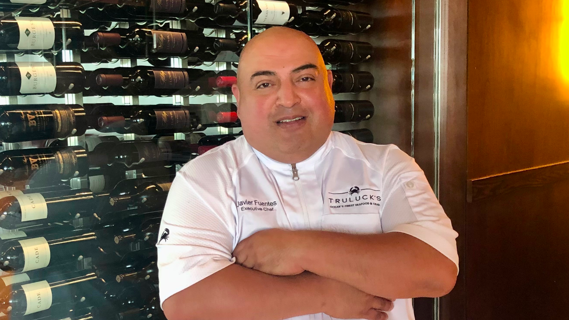 chef Javier Fuentes of Truluck's Rosemont in a white logoed chef coat standing in front of the wine wall with his arms folded across his chest.