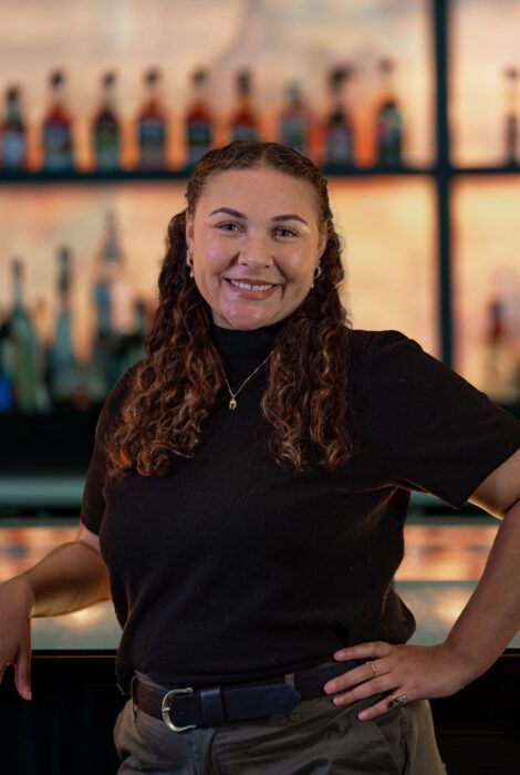 headshot of Kyla Adams, sales and events manager for Truluck's Houston, standing in front of the bar top with one hand on her hip and an elbow on the bartop, she's smiling and wearing a black blouse