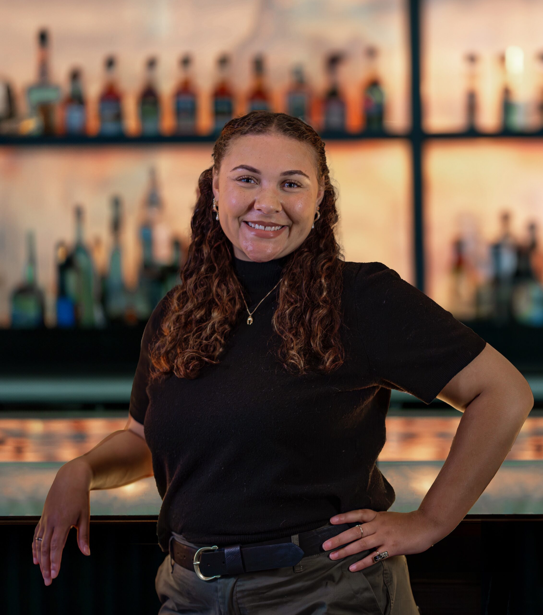 headshot of Kyla Adams, sales and events manager for Truluck's Houston, standing in front of the bar top with one hand on her hip and an elbow on the bartop, she's smiling and wearing a black blouse