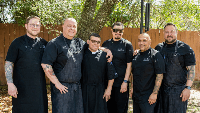 Chef team standing together outside at Camp Hope all are wearing black chef coats and aprons.