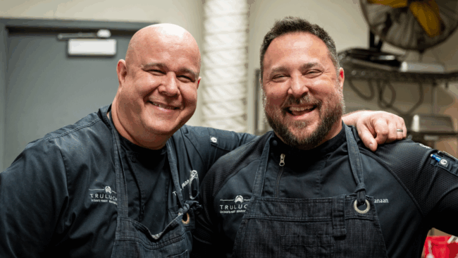Chef Thomas and Chef Sammy smiling together with Chef Thomas' arm on Sammy's shoulder. They are wearing black chef coats and black denim aprons