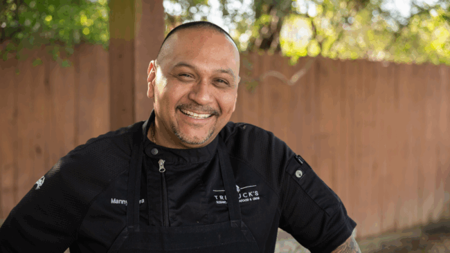Chef Manny outside on the porch at Camp Hope wearing a black chef's coat and black apron