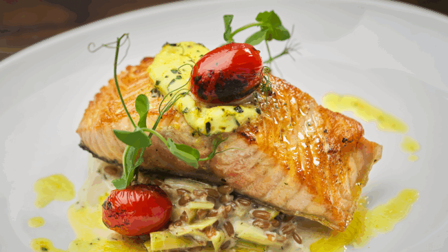 overhead view of Ora King Salmon filet with blistered tomatoes, fresh pea tendrils, farro & leek fondue, citrus-tarragon butter on a white china plate