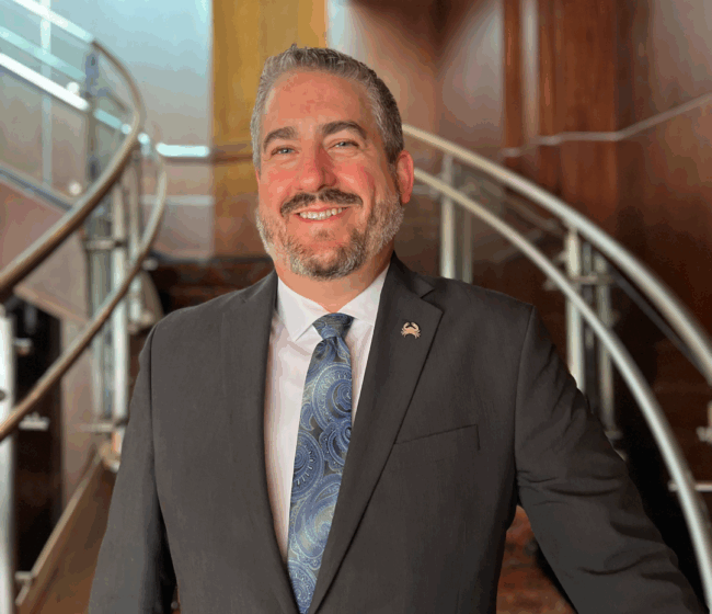 headshot of Ryan Studt, general manager of Truluck's Dallas, wearing a suit and tie and standing at the bottom of the staircase at Truluck's Dallas location