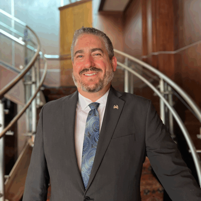 headshot of Ryan Studt, general manager of Truluck's Dallas, wearing a suit and tie and standing at the bottom of the staircase at Truluck's Dallas location
