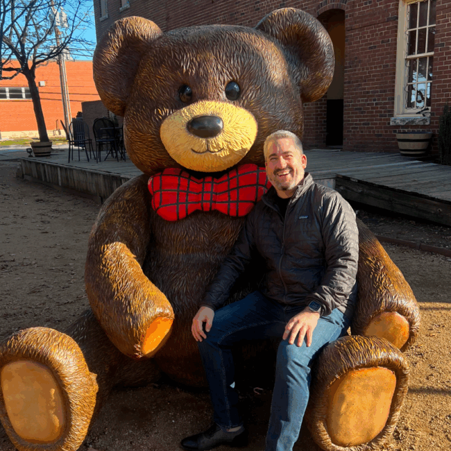 Ryan Studt smiling and sitting on a giant teddy bear statue outside of a building