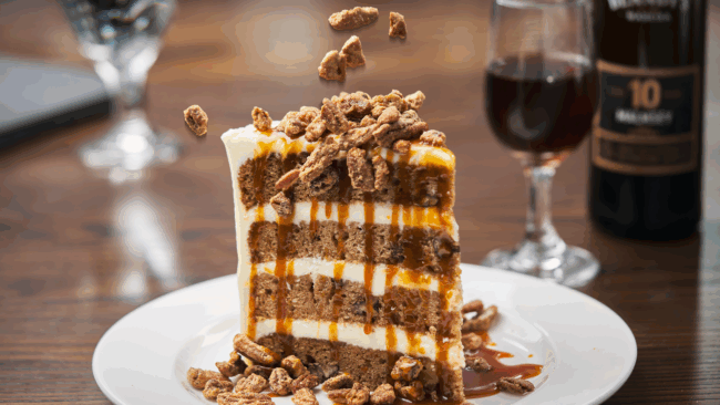 4 layers of carrot cake with cream cheese icing drizzled with caramel sauce and spiced pecans being dropped onto the cake