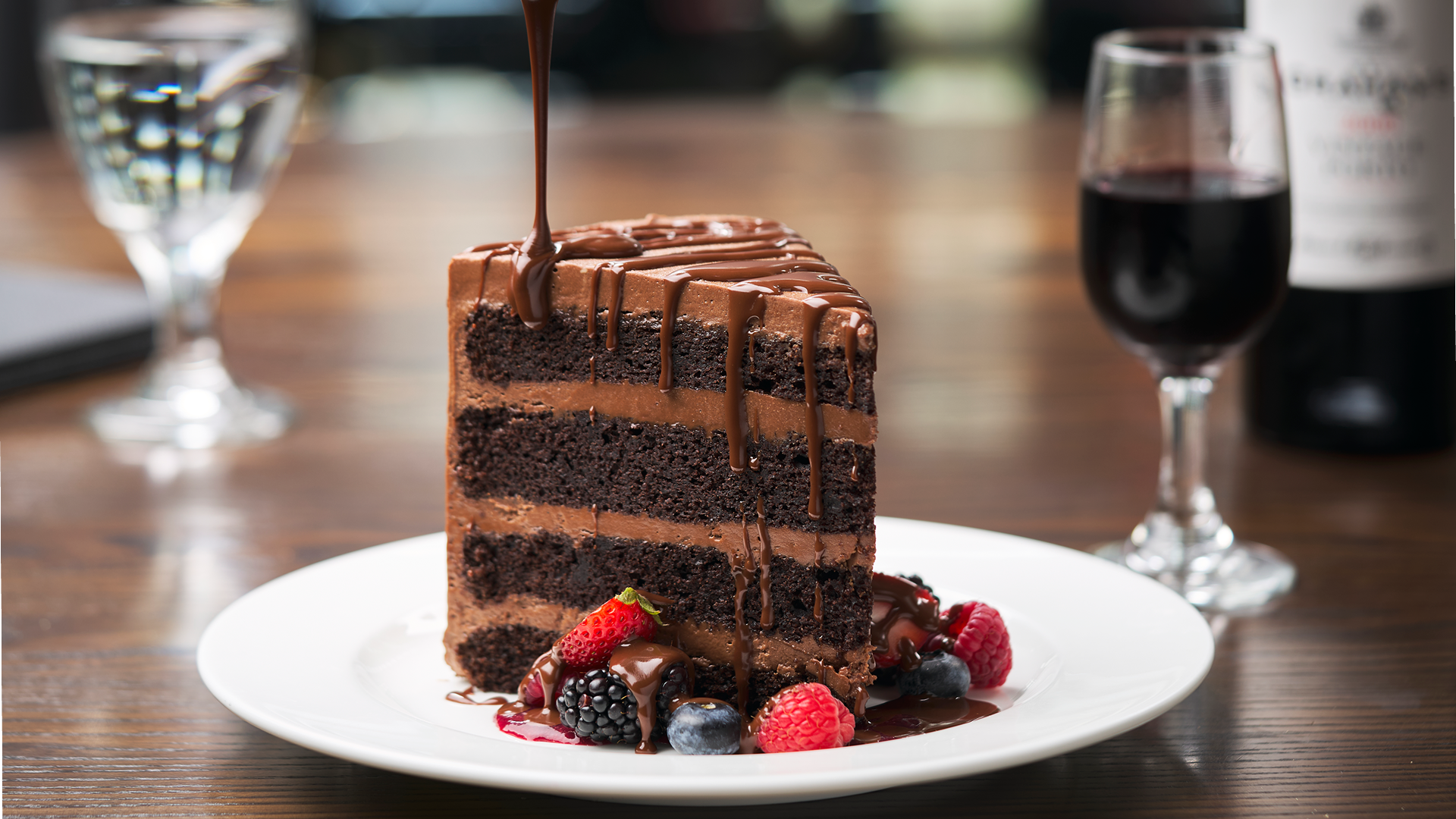 4 layer chocolate cake with chocolate icing on a white plate with fresh berries at the base of the cake and chocolate ganache being drizzled over the top and down the sides of the cake