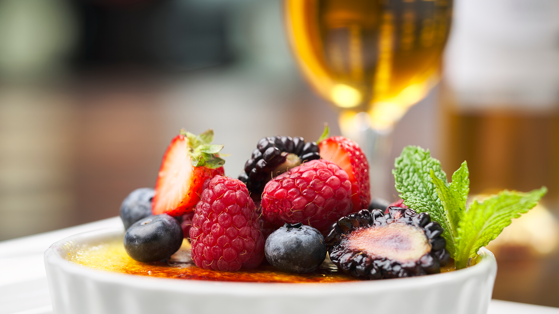 Close up image of fresh raspberries, blackberries, strawberries and blueberries on top of a creme brulee dessert in a white ceramic dish with a fresh mint garnish