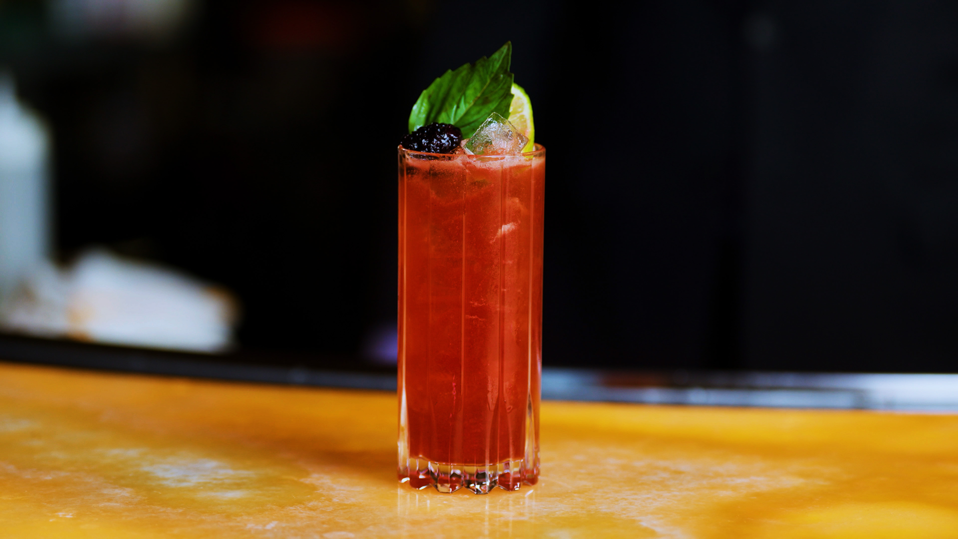 A non alcoholic tall, vibrant red hibiscus fresca served over ice in a fluted highball glass, garnished with fresh mint leaves, a blackberry, and a lime wedge, set on a polished bar with a softly blurred background.
