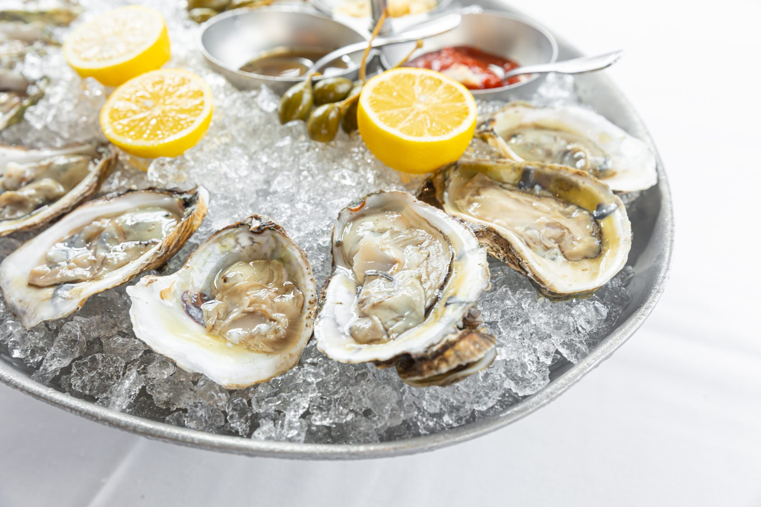 fresh shucked oysters on a platter of crushed ice with fresh lemon halves, caper berries and sauces