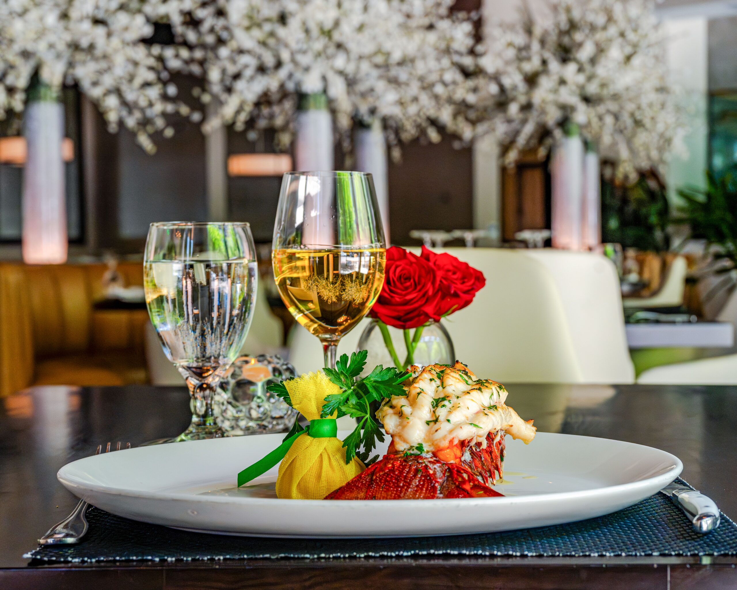 A plated broiled lobster tail garnished with herbs and served with a wrapped lemon sits on a white dish beside a glass of white wine and sparkling water in Truluck’s upscale dining room, with red roses and floral décor in the background.