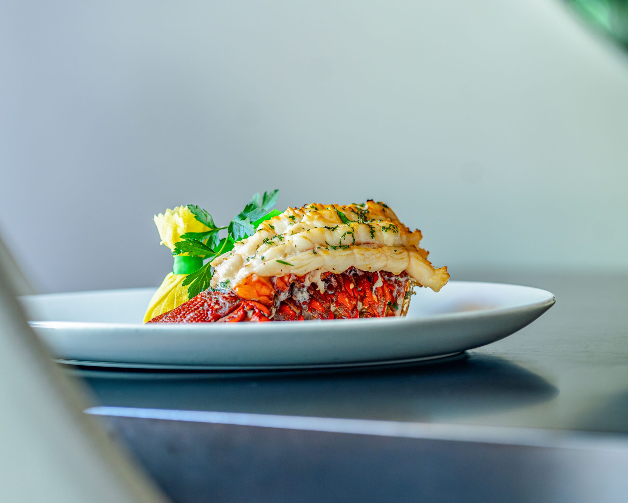 A broiled lobster tail topped with herbs sits on a white plate with a wrapped lemon and fresh parsley garnish.