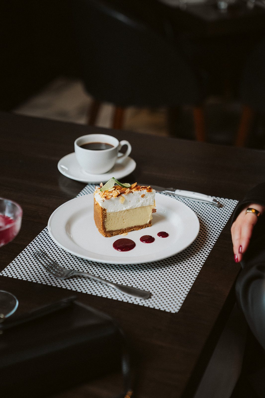 a slice of key lime pie on a white china plate with 3 dollops of raspberry sauce and a cup of coffee in the background