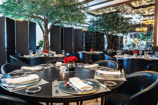 Elegant group dining room at Truluck’s featuring round tables set with polished glassware, navy chairs, red rose centerpieces, and indoor olive trees against mirrored walls.