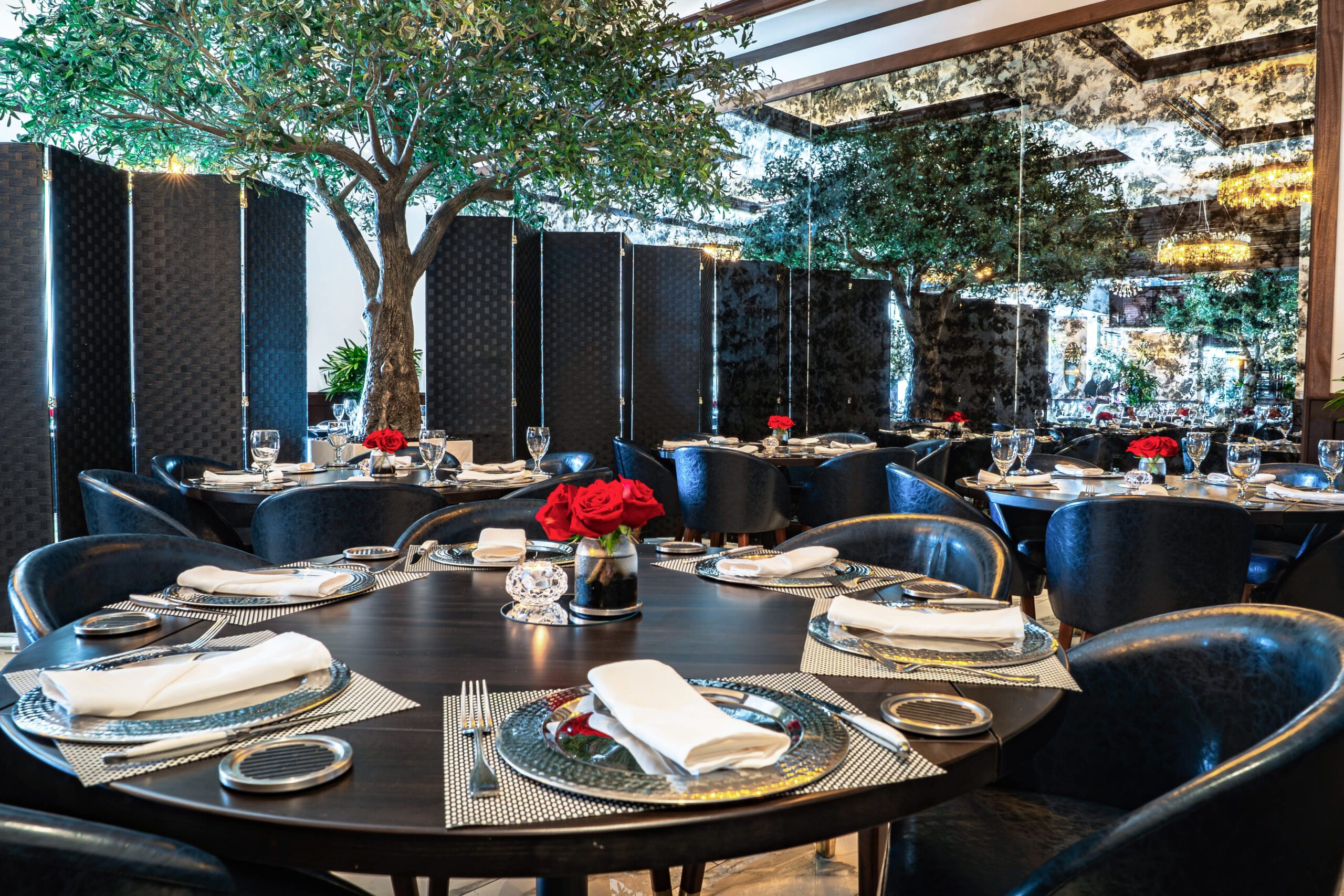 Elegant group dining room at Truluck’s featuring round tables set with polished glassware, navy chairs, red rose centerpieces, and indoor olive trees against mirrored walls.