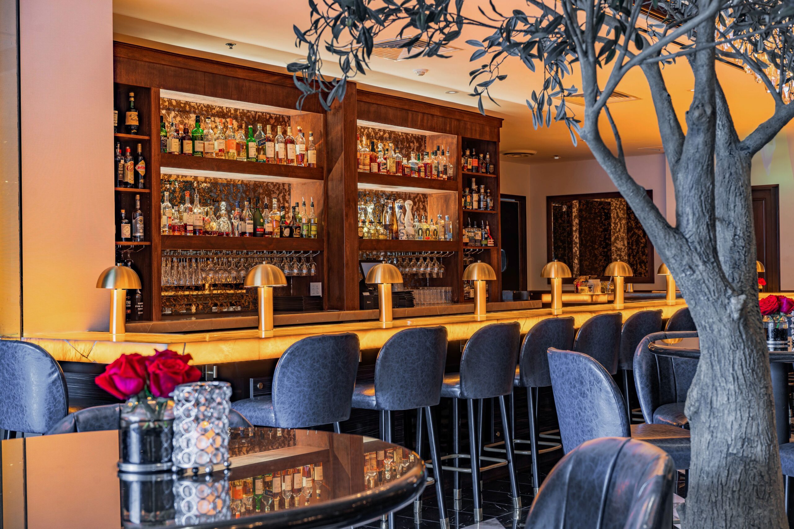 Warm, upscale Truluck’s bar illuminated by a glowing onyx counter, crystal chandeliers, and gold accents, with navy leather seating and marble floors creating a sophisticated lounge setting.