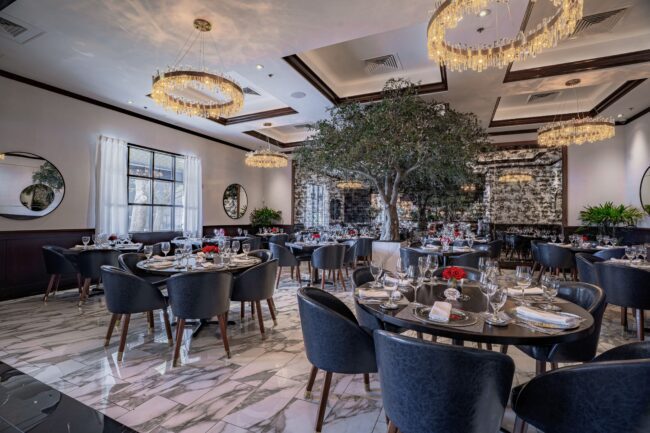 A refined group dining restaurant interior featuring dark round tables, navy leather seating, gleaming marble floors, cascading crystal chandeliers, and a centerpiece indoor tree that anchors the space. Red rose arrangements and round wall mirrors complete the luxurious design.