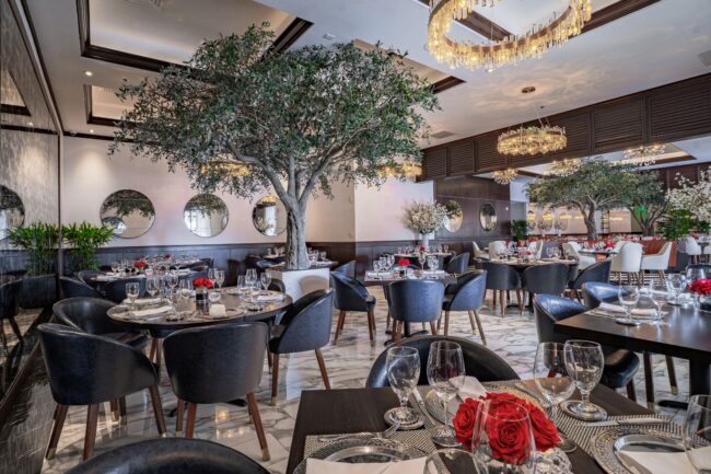 Upscale TRU group dining room with round tables, navy leather chairs, marble floors, crystal chandeliers, and large indoor trees.