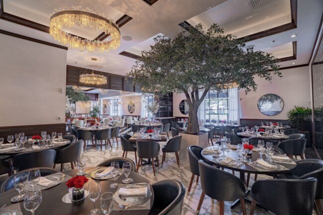 An elegant dining room with round tables set with polished glassware, white napkins, and red rose centerpieces. Navy leather chairs surround the tables, while marble floors, large crystal chandeliers, indoor trees, and round mirrors create a sophisticated, nature-inspired atmosphere.