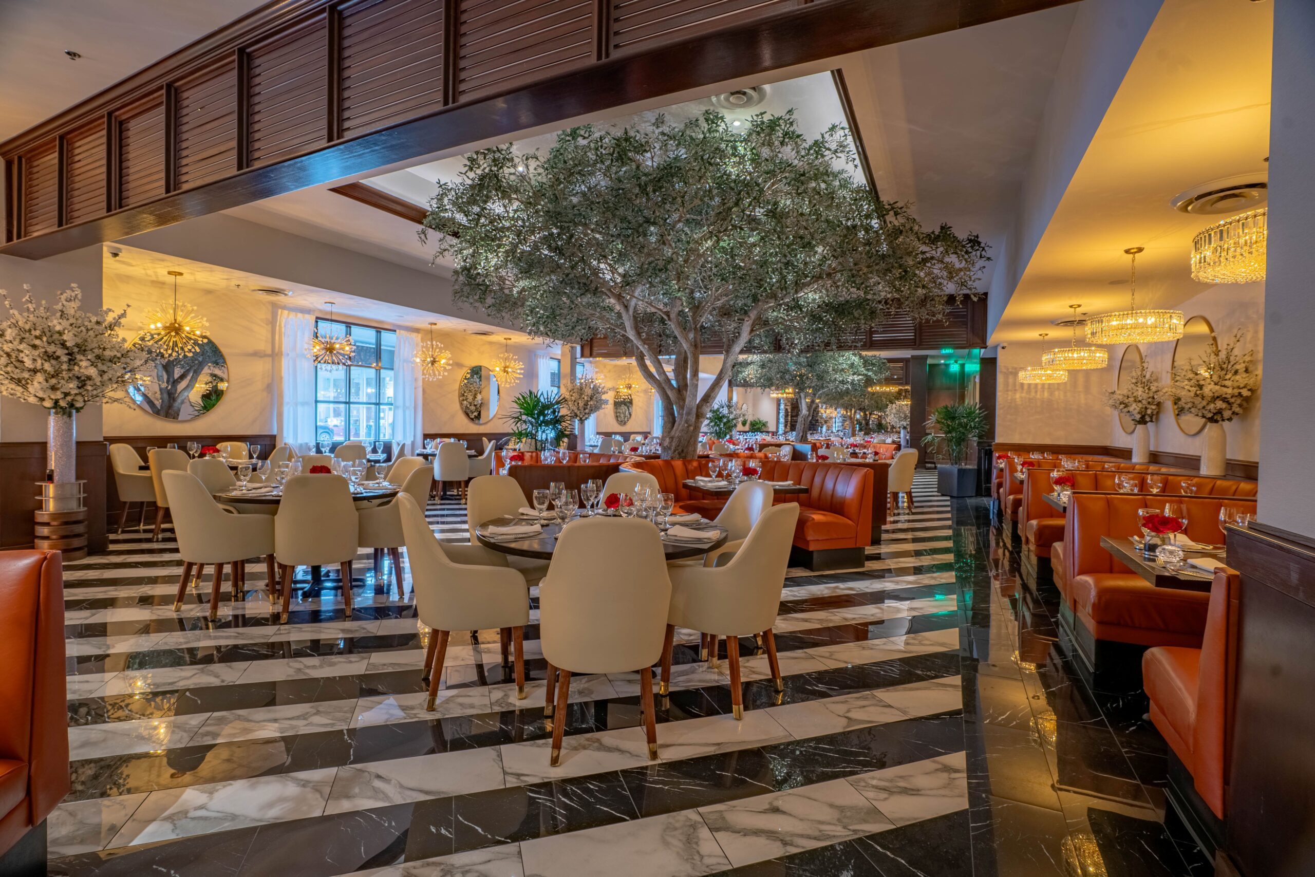 A beautifully appointed dining area with tables set for guests, surrounded by cream chairs, marble floors, statement chandeliers, floral arrangements, and large mirrors that amplify the warm, luxurious feel of the restaurant.