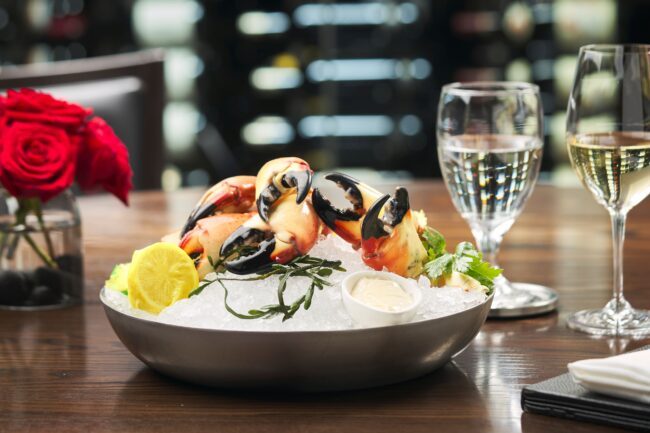 a platter of fresh Florida stone crab claws on a bed of crushed ice with fresh seaweed, a wrapped lemon and a small ramekin of mustard sauce sitting on a table with a glass of wine and fresh red roses in the background