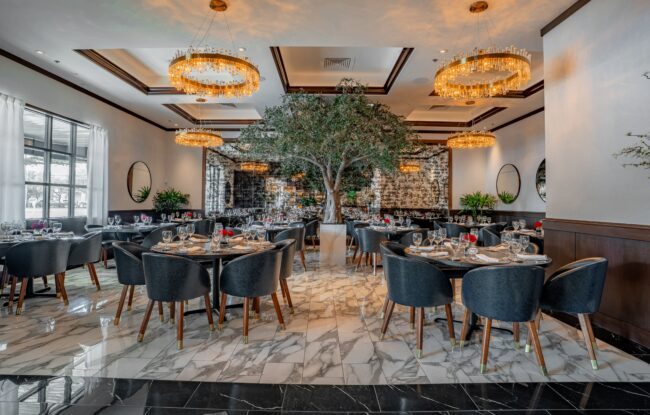 Upscale Truluck’s Houston TRU group dining room featuring navy leather chairs, round tables, marble flooring, gold ring chandeliers, mirrored walls, and a large olive tree at the center.