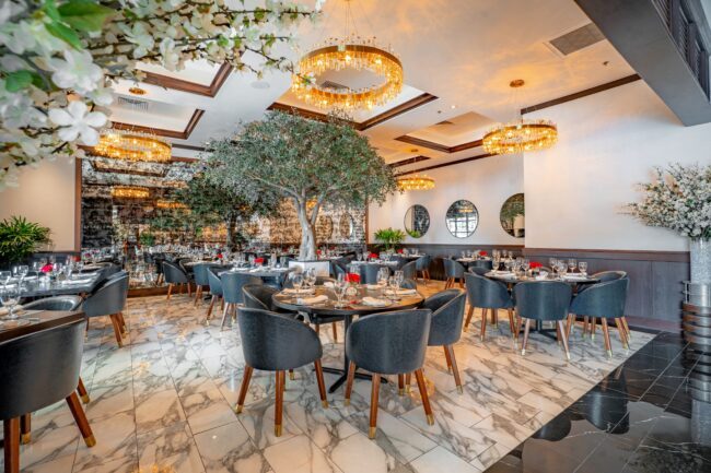 Sophisticated Truluck’s Houston TRU group dining room with navy chairs, marble floors, gold chandeliers, and a large olive tree centerpiece.