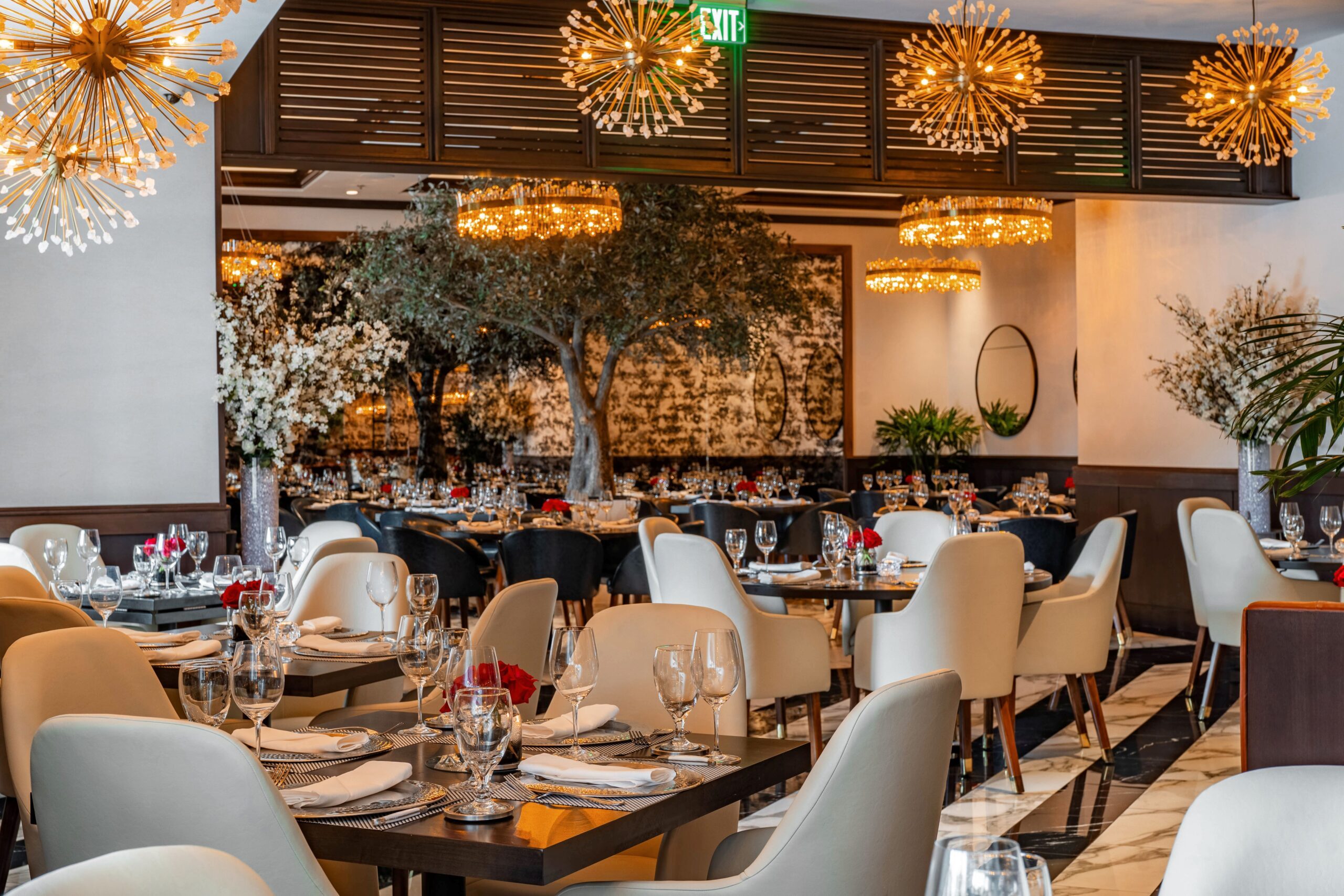 Elegant Truluck’s dining room with cream chairs, black tables, gold starburst chandeliers, and a large central olive tree.