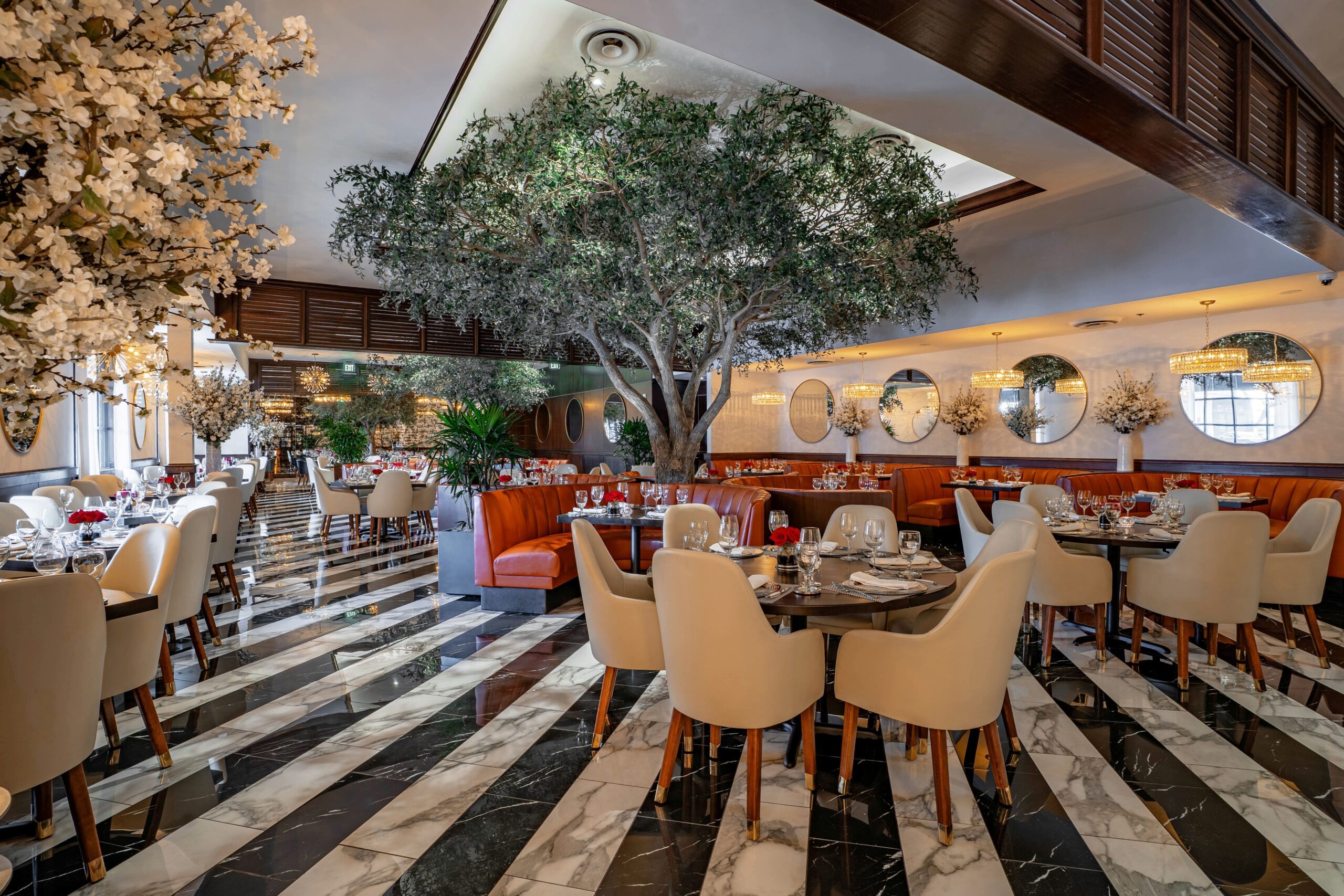 Bright, upscale Truluck’s dining room featuring cream upholstered chairs, orange leather booths, a large olive tree centerpiece, round mirrors, and black-and-white marble flooring.