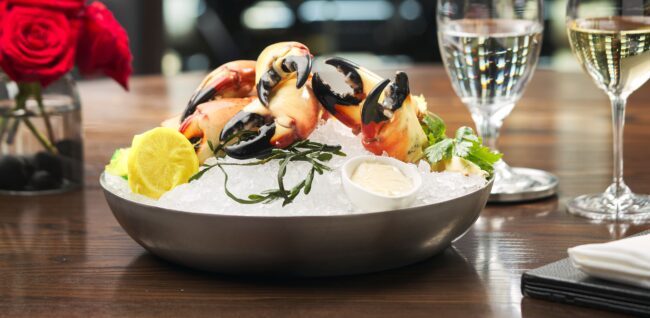 Fresh Florida stone crab claws served chilled on crushed ice in a silver bowl, garnished with lemon, seaweed, and parsley, alongside a ramekin of creamy mustard sauce, with Champagne glasses and red roses softly blurred in the background.