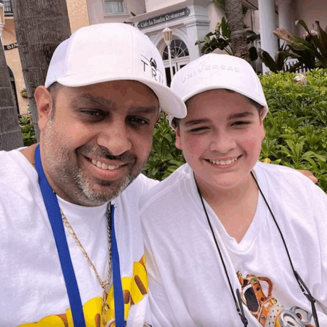 Blake Goldie smiling alongside his son outdoors, both wearing white hats and enjoying a sunny day near a restaurant