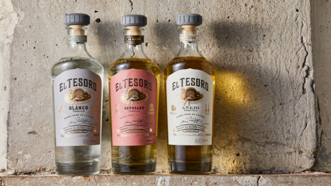 El Tesoro Blanco, Reposado, and Añejo tequila bottles displayed side by side with warm light reflecting on a stone background.