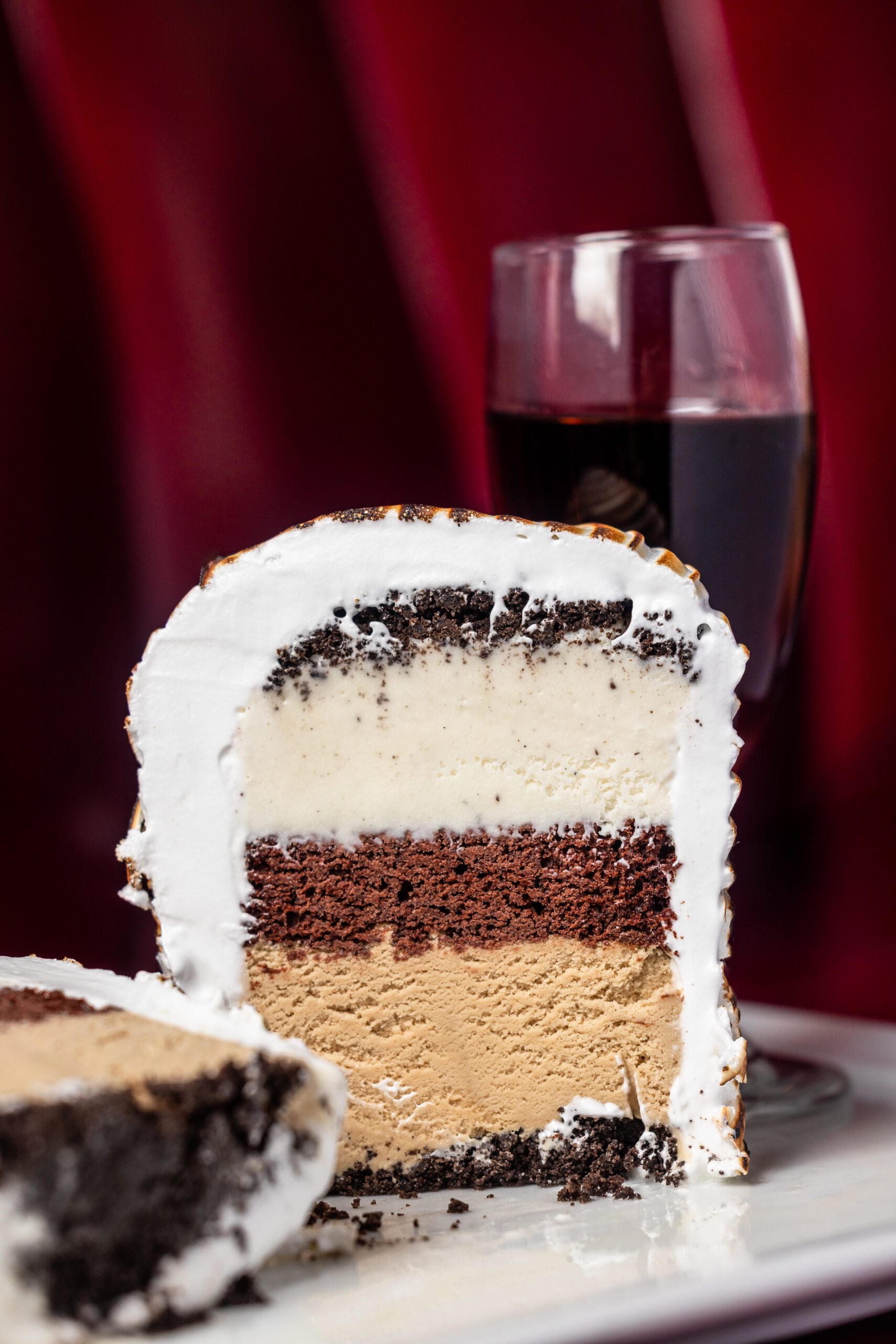 A sliced baked Alaska dessert showing distinct layers of chocolate cake, coffee-colored ice cream, and vanilla ice cream, encased in toasted meringue, plated with a glass of red wine against a rich red backdrop.