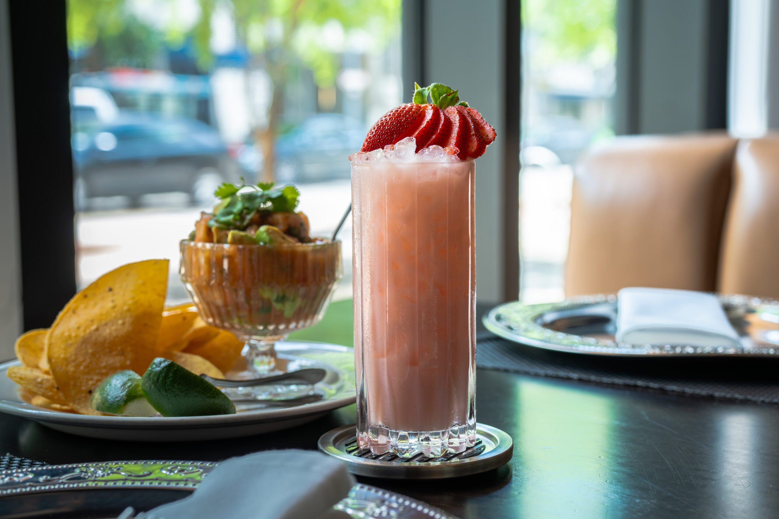 Berry Patch non-alcoholic cocktail served over ice in a tall glass, garnished with fresh sliced strawberries, set beside chips and campechana on a dark dining table with natural light from nearby windows.