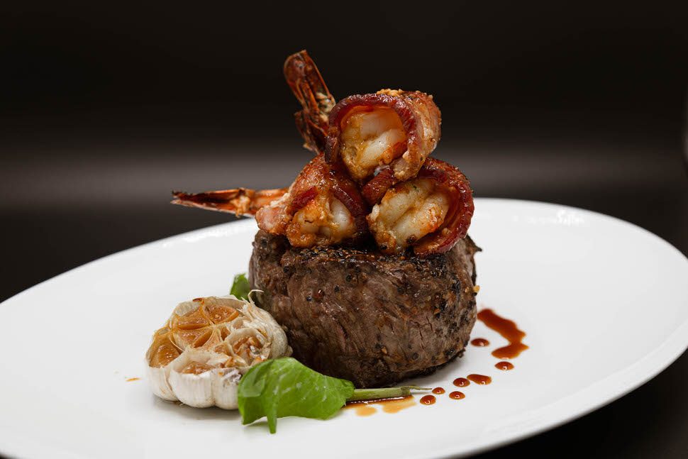 12 oz filet mignon topped with cherrywood bacon-wrapped shrimp, served on a white plate with roasted garlic, a fresh basil leaf, and a drizzle of steak sauce.