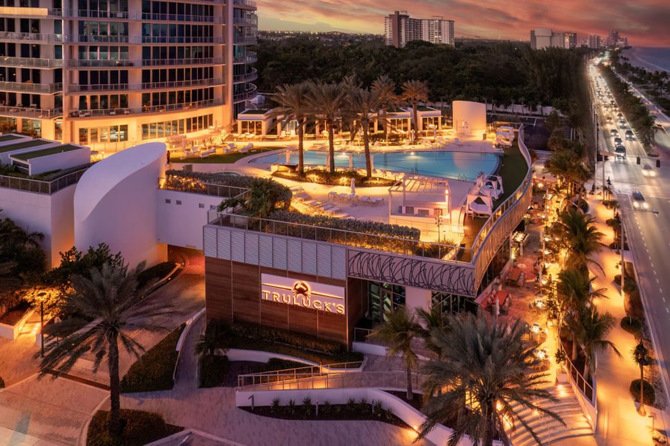 Aerial sunset view of Truluck’s Fort Lauderdale along the beachfront, featuring the illuminated Truluck’s sign, rooftop pool and palm trees at the Paramount Residences, and the glowing coastline stretching into the distance.