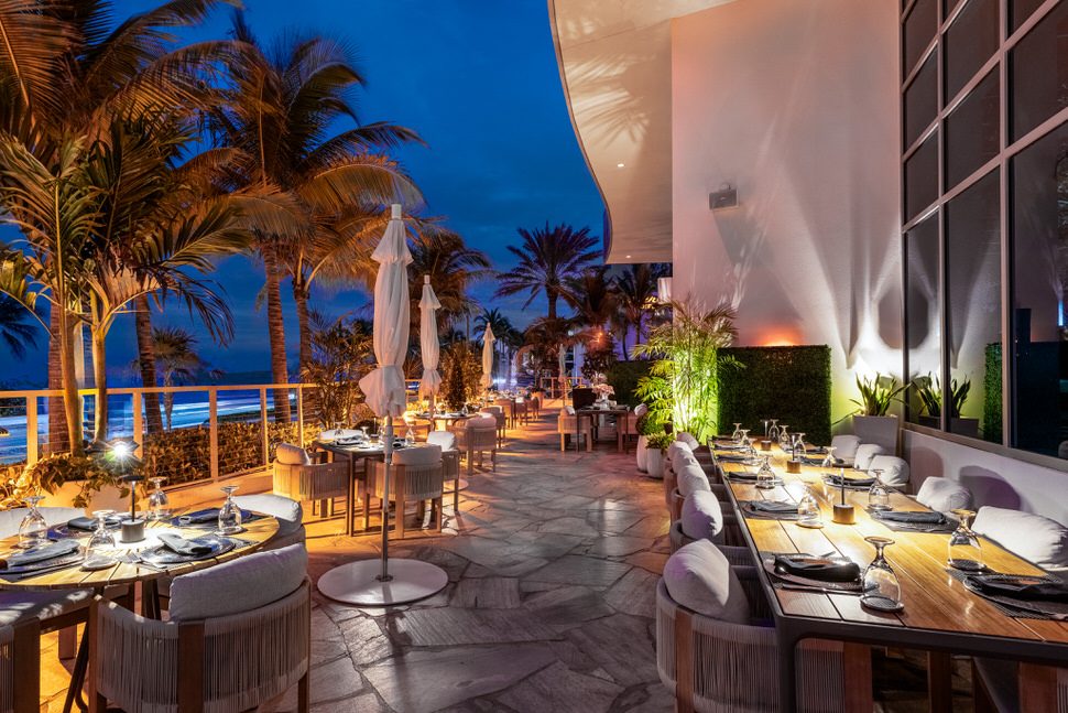 Oceanfront patio at Truluck’s Fort Lauderdale at dusk, featuring palm trees, softly lit tables set with glassware and linens, white umbrellas, and sweeping beach views along the coastline.