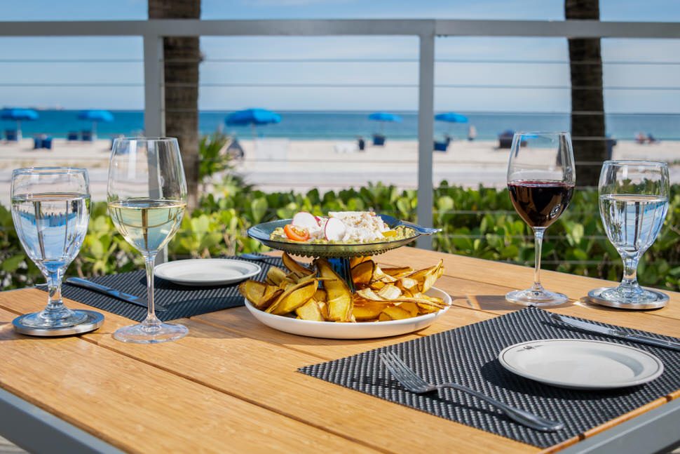 Beachfront patio dining at Truluck’s Fort Lauderdale, featuring a wooden table set with wine and water glasses, a seafood appetizer with house-made chips, and ocean views with palm trees and blue umbrellas in the background.