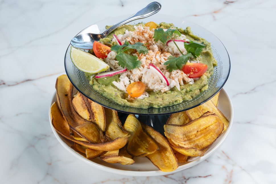 Truluck’s Fort Lauderdale blue crab guacamole topped with fresh lump crab, cherry tomatoes, radish slices, cilantro, and a lime wedge, served with house-made plantain chips on a white plate over a marble surface.